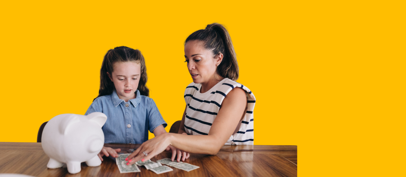A mother sits at a table with her daughter, teaching her how to count money from a piggy bank.