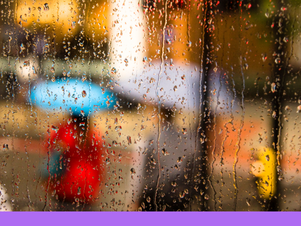 Rain falling on a window with people holding umbrellas blurred through the window. 